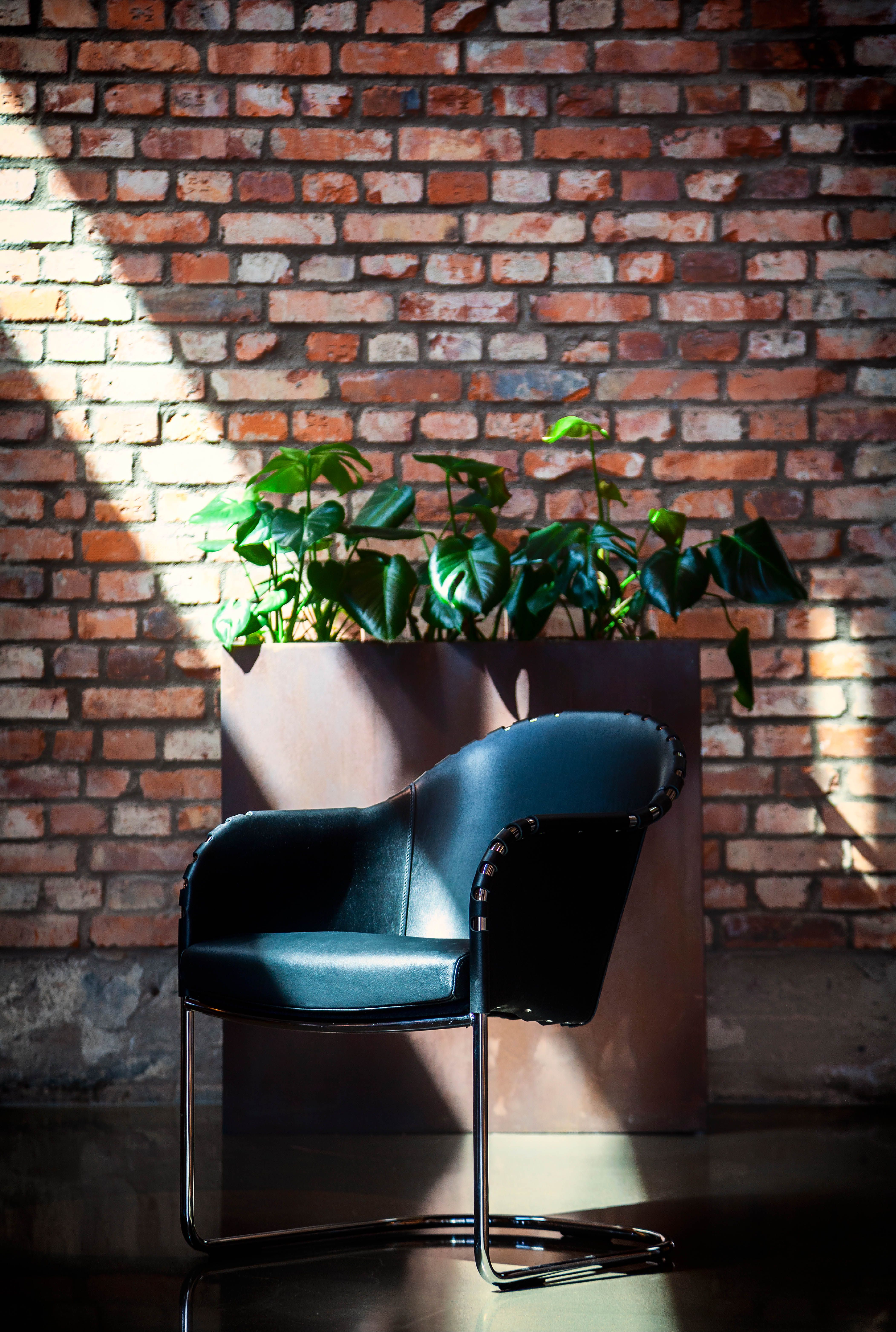 Ingo armchair in dark leather in front of a brick wall, indoors.